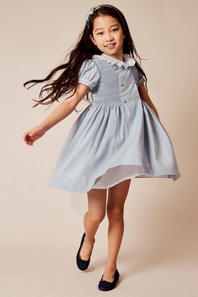 a fullbody studio shot of a young girl with long brown hair wearing a beautiful light blue smocked dress with a peter pan collar and white slip underneath.  she's smiling and playfully moving, wearing black ballet flats and a hair bow. the background is a soft beige.  the overall impression is one of sweetness and childhood innocence.