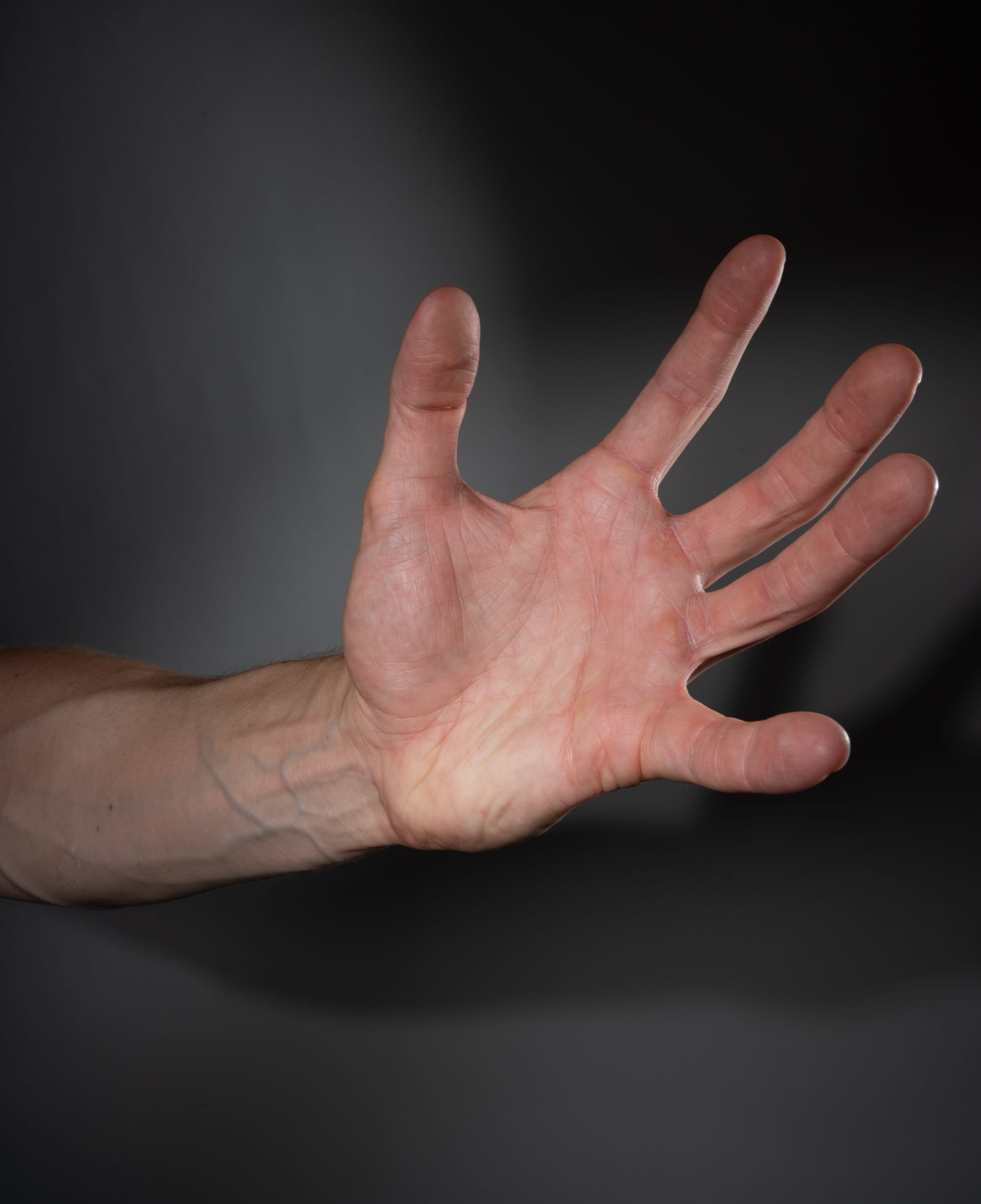 a closeup image of a man's outstretched hand set against a dark gray background. the hand is welllit, revealing prominent veins and the texture of his skin.  the fingers are slightly spread, as if reaching out or gesturing.