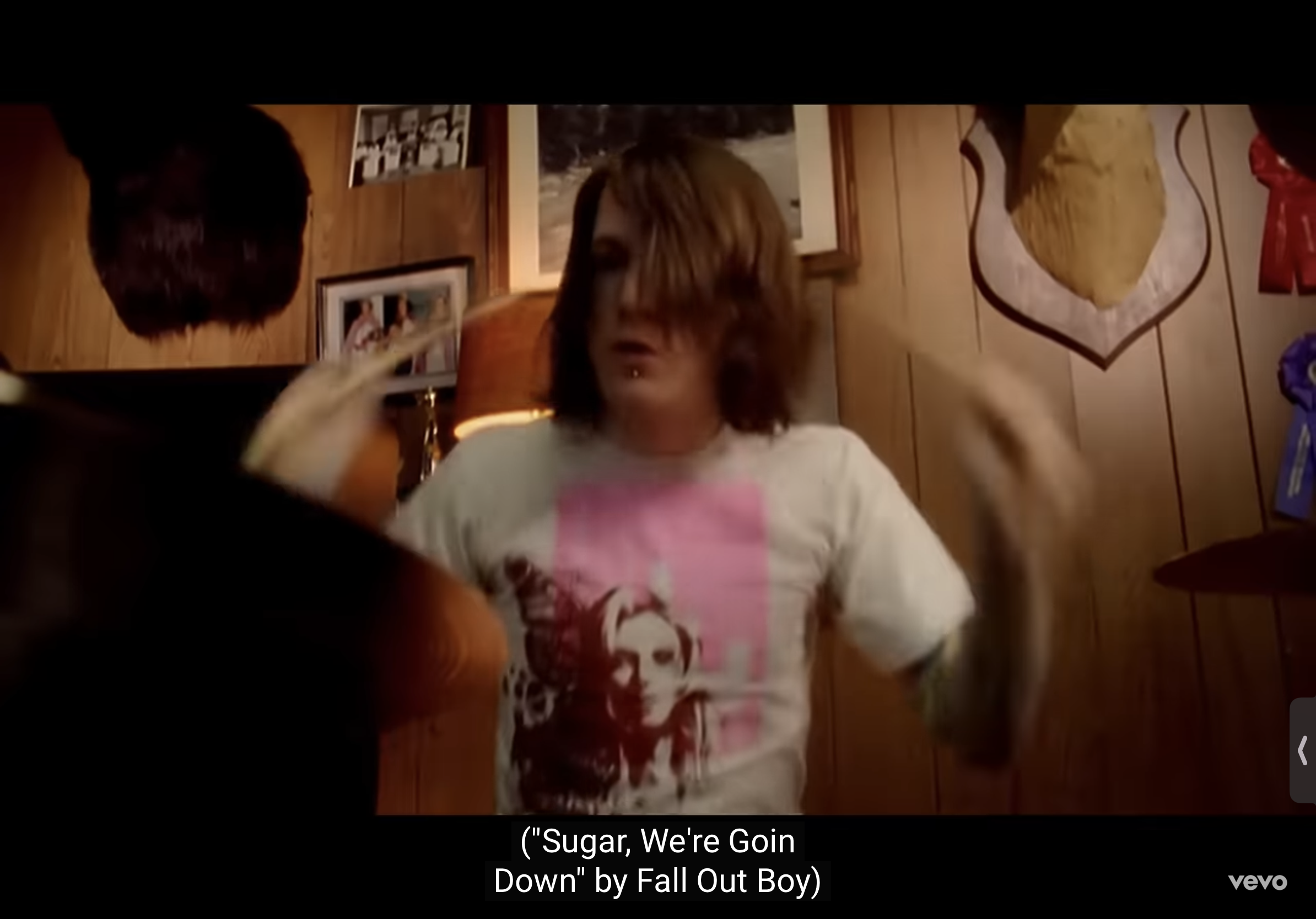 a man with shoulder length brown hair and wearing a white tshirt plays the drums.  he is looking directly at the camera and smiling with his mouth open. the wall behind him is wood paneled and features framed photos and a mounted deer head.