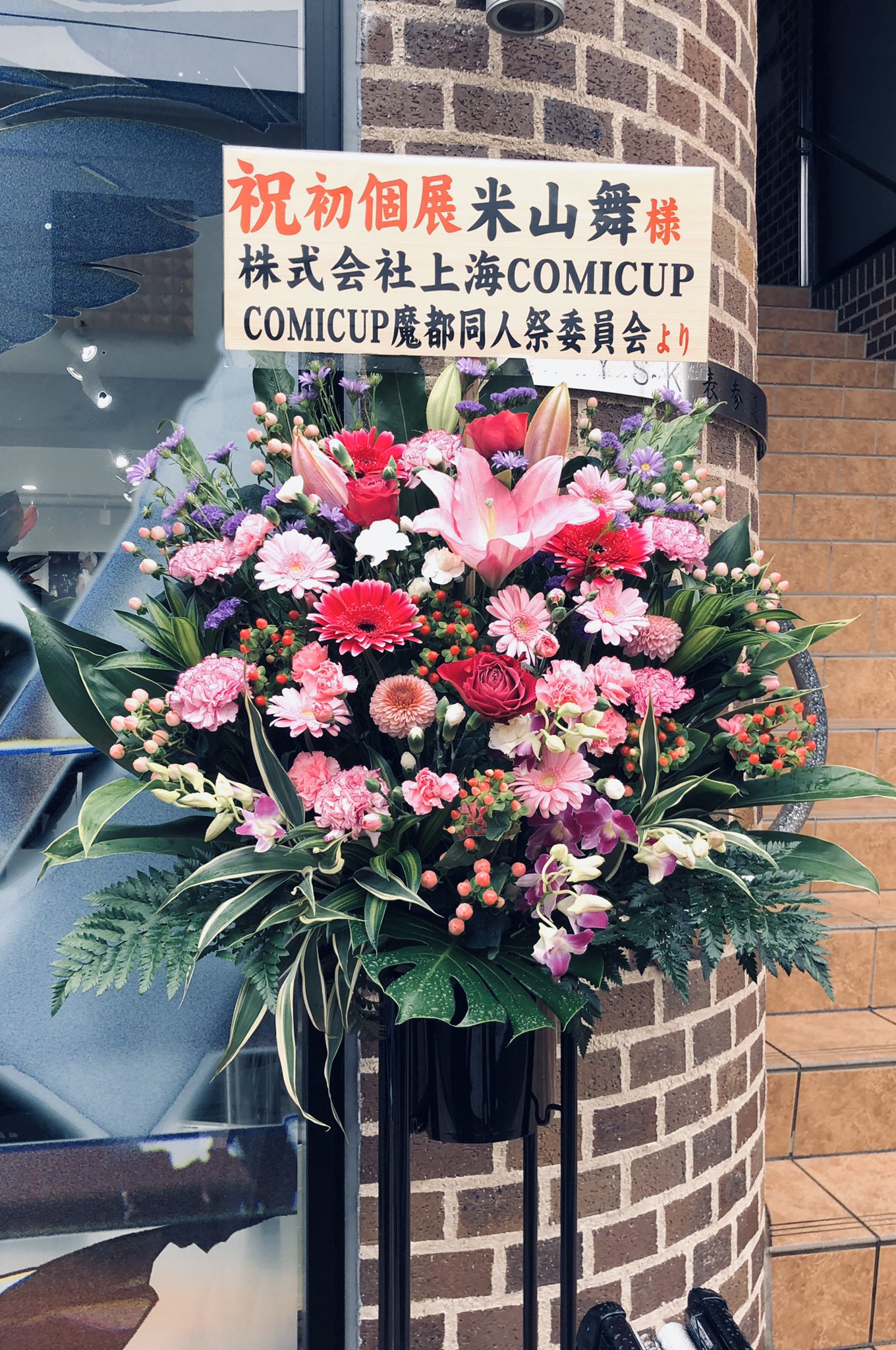 a colorful flower arrangement standing in front of a brick wall. there is a sign on the wall with japanese text that congratulates someone on their first solo exhibition.```