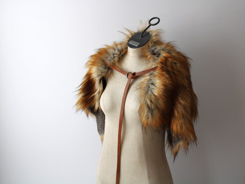 a brown and orange faux fur stole is displayed on a mannequin. the stole has a brown leather strap that goes around the neck and down the front. the mannequin is against a white background.