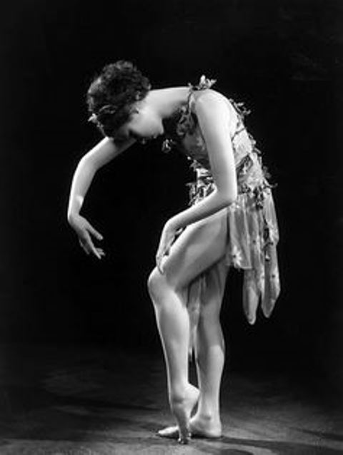 the image is a striking black and white photograph of a ballerina in a flowing costume. she is posed in a graceful, almost pensive bend, her body curved in a fluid line. her legs are slender and elegantly positioned, her feet delicately poised. the dancer's costume appears light and airy, possibly made from silk or a similar material, with folds that catch the light. the background is dark and uncluttered, allowing the ballerina to stand out as the central focus. the overall aesthetic is both classic and artistic, suggesting a vintage style, perhaps from the 1920s or 1930s. the mood is contemplative and serene.