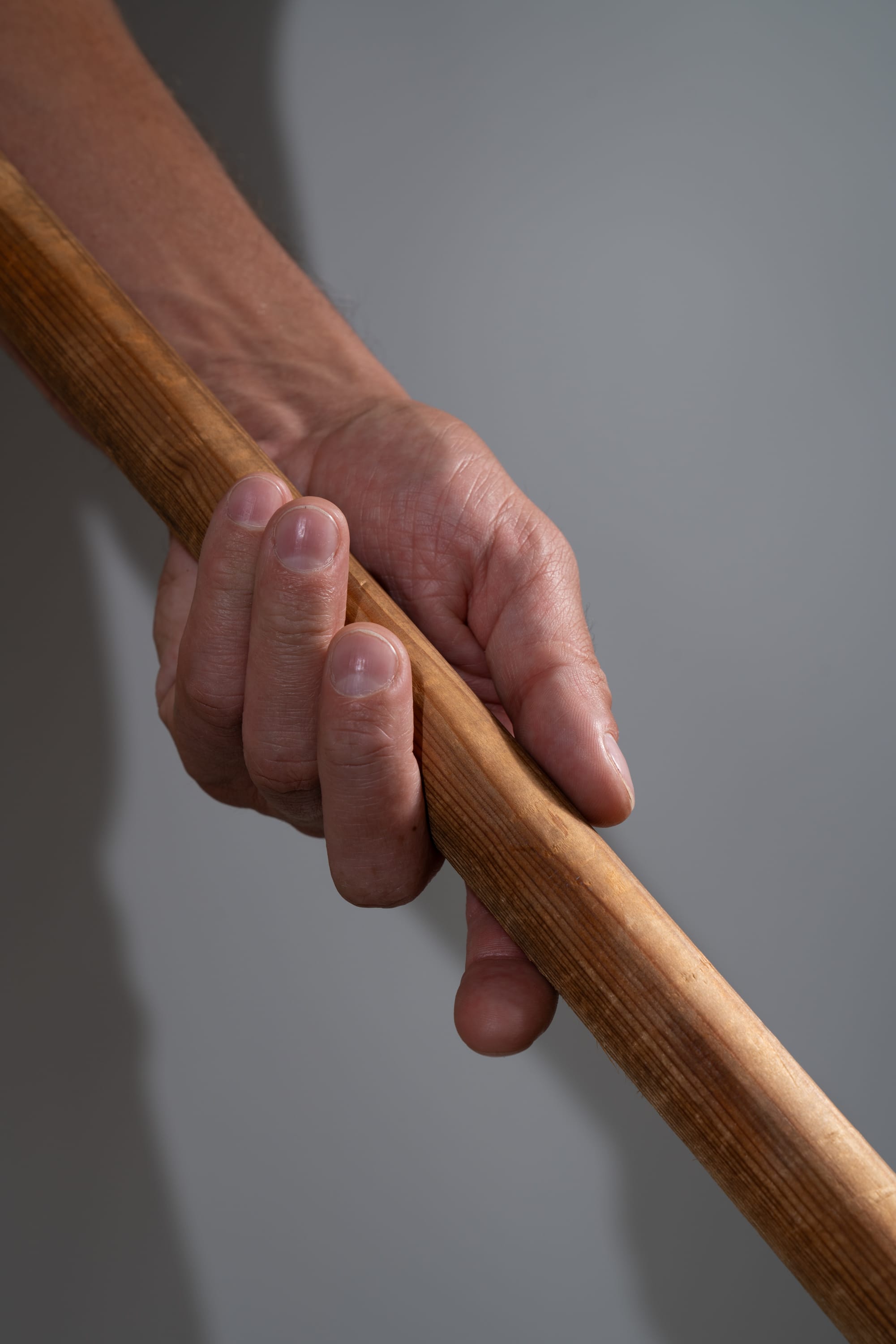 a closeup view of a man's hand firmly gripping a wooden stick against a gray background. the artificial lighting casts a shadow, emphasizing the strength and tension in the hand.