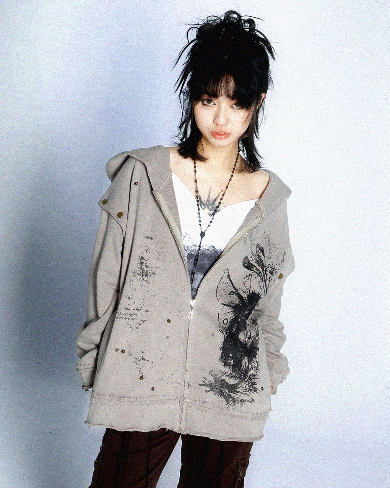 the image shows a young woman with short, dark hair styled in a choppy, slightly grunge manner. she is wearing an oversized, light grey zipup hoodie adorned with a distressed, worn look, and features a dark graphic print of what appears to be a moth or similar insect.  the hoodie is loosely fitted and has a raw, unfinished hem.  the model also wears a simple white shirt underneath and a black necklace. she has visible tattoos on her neck and chest.  the overall style leans toward grunge, alternative, or dark academia aesthetics. the background is plain and lightcolored, providing a neutral backdrop that emphasizes the clothing. the image appears to be a studio shot, likely for a fashion catalog or online store.