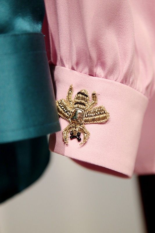 the image is a closeup shot focusing on the sleeve cuffs of two women's shirts. one shirt is a soft pink and features a detailed gold and black beaded bee embroidered on the cuff. the other shirt, partially visible, is a deep teal color. the image highlights the fine craftsmanship and luxurious details of the pink shirt's embroidery.  the contrast between the colors and textures of the two shirts is also visually striking.
