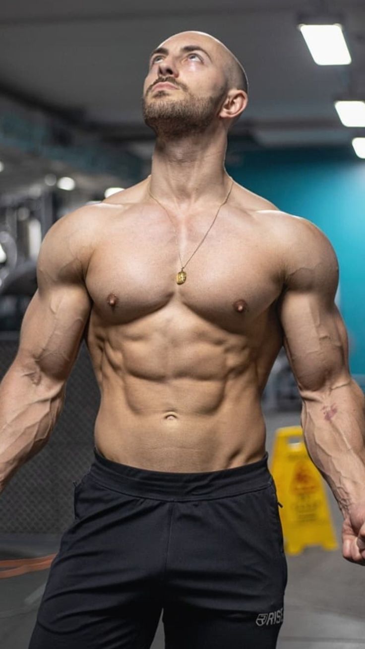 a shirtless man with a very defined sixpack and muscular physique stands in a gym setting, looking up at the ceiling. he is wearing black athletic shorts with a small logo on the right leg, and he has a gold chain around his neck. he is looking up at the ceiling with a determined expression on his face, suggesting he is about to begin a workout.