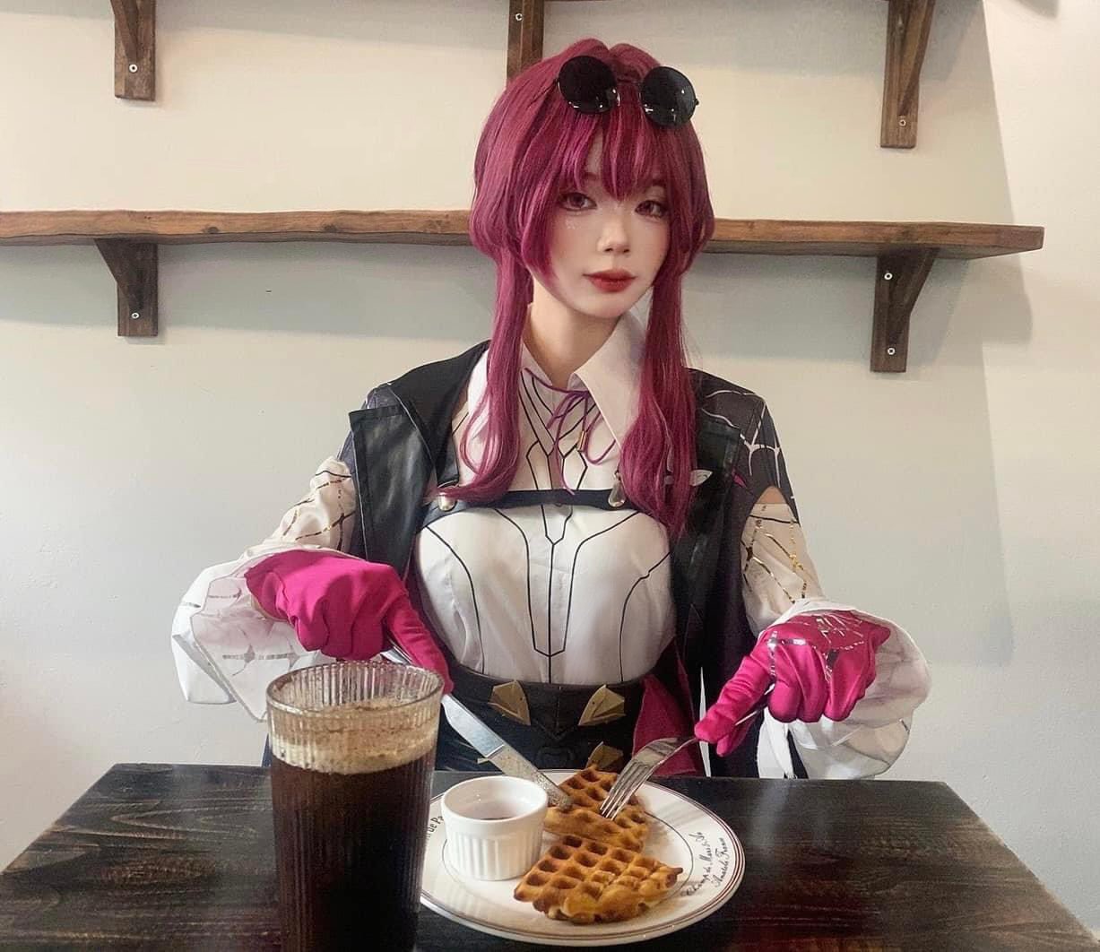 a woman with pink hair, wearing a black coat, a white shirt, sunglasses, black gloves, and pink gloves, is sitting at a table in a restaurant, eating a waffle with a fork. she is also drinking coffee from a glass. the table has a wooden surface and the walls are white. there is a shelf on the wall behind the woman.