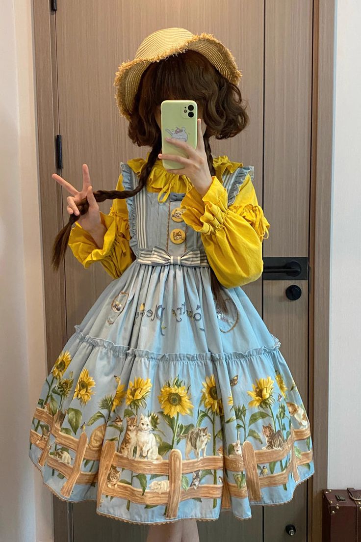 a person is taking a mirror selfie wearing a blue lolita fashion dress. the dress has sunflowers and cats on it. they are also wearing a yellow blouse and a straw sun hat.