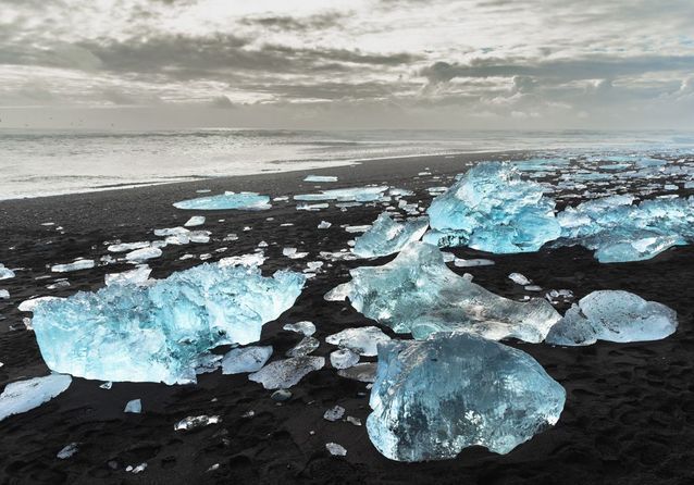 the image shows a black sand beach in iceland, covered with chunks of ice and icebergs. in the background there is the sea and the sky with the clouds. the scene is at diamond beach.