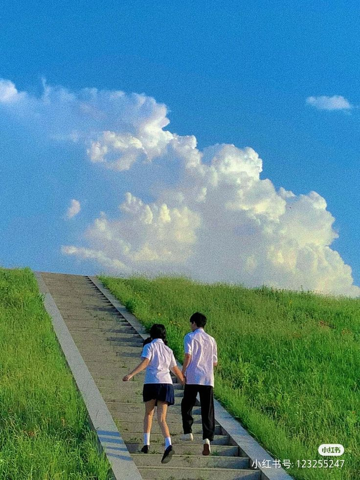 a young couple is walking up a long flight of concrete stairs leading up a grassy hill under a bright blue sky with fluffy white clouds. the girl is wearing a white collared shirt and a short blue skirt, while the boy is wearing a white collared shirt and black pants. they are holding hands as they ascend the stairs, suggesting a sense of companionship and connection. the overall scene is peaceful and suggests a romantic or leisurely moment in nature.