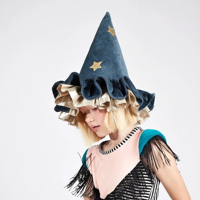 a young girl with blond hair is wearing a blue wizard hat with gold star embellishments and a ruffled brim. she is looking down and slightly to the left. she is wearing a pink and black striped dress with teal and black fringe detailing. the photo was taken in a studio against a white background.