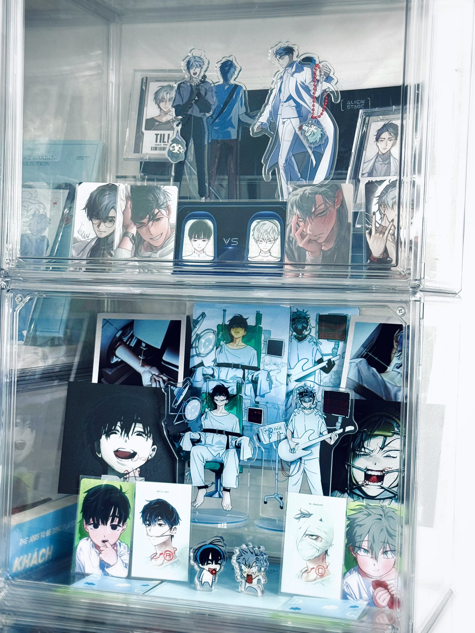 a closeup shot of a clear acrylic display case filled with anime figures and artwork. the case is divided into two shelves. the top shelf has several figures of characters from different anime series. the bottom shelf has a collection of images and smaller figurines, primarily featuring a group of male characters in white medical attire. the overall aesthetic is clean and minimalist, showcasing the collection with care.