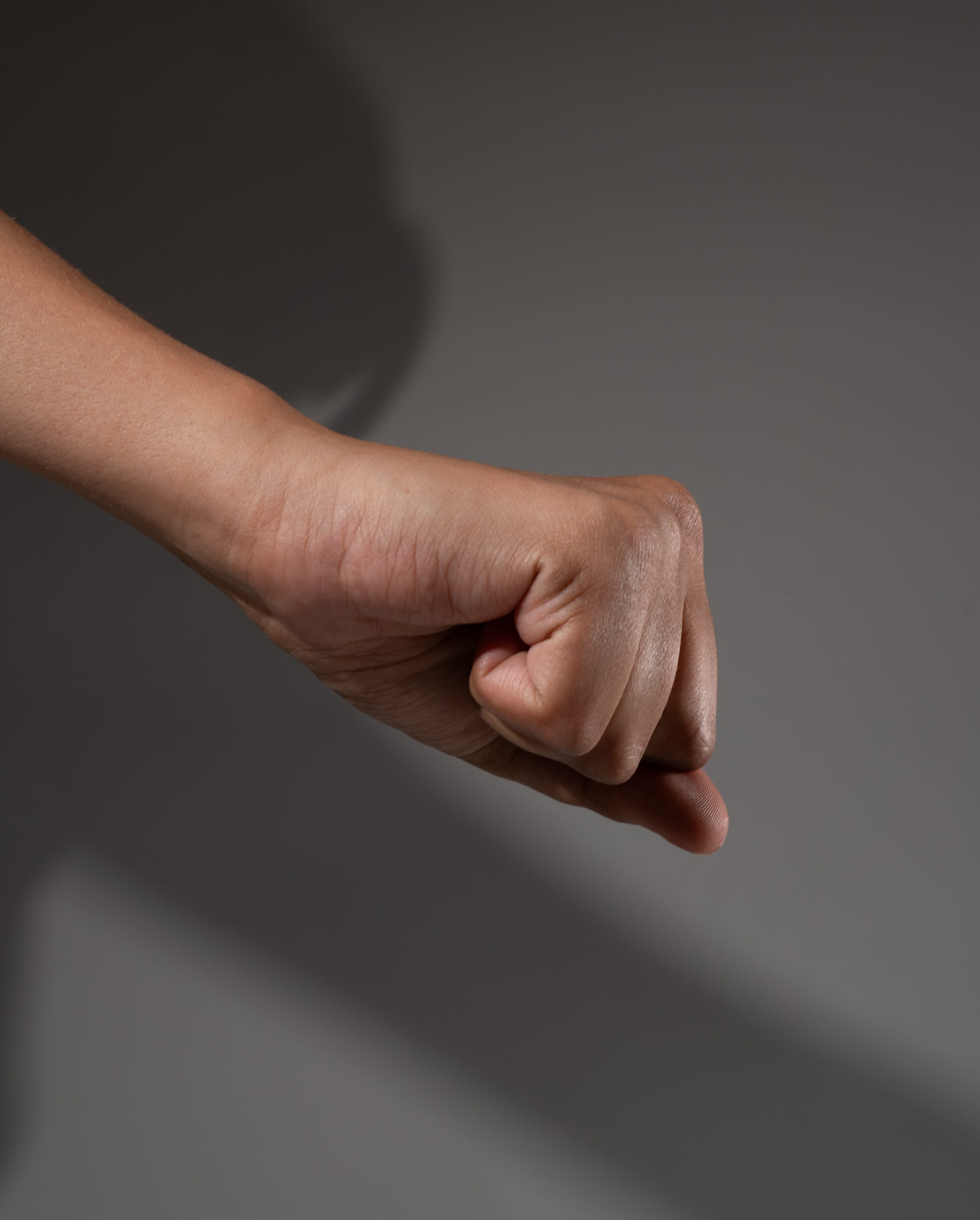 a clenched fist is held up against a gray background, casting a long shadow. the hand is illuminated by strong studio lighting, highlighting the details of the skin and the tension in the muscles. the image evokes a sense of power, determination, and resilience.