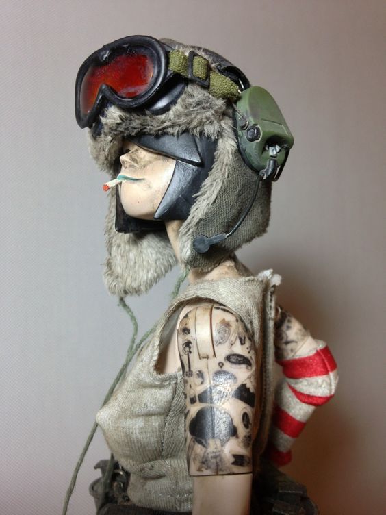 closeup of an action figure of a female soldier with a helmet, goggles, and gloves. she's wearing a tattered military uniform and has tattos all over her arms. she appears to be holding a cigarette in her mouth. the figure is isolated on a white background.