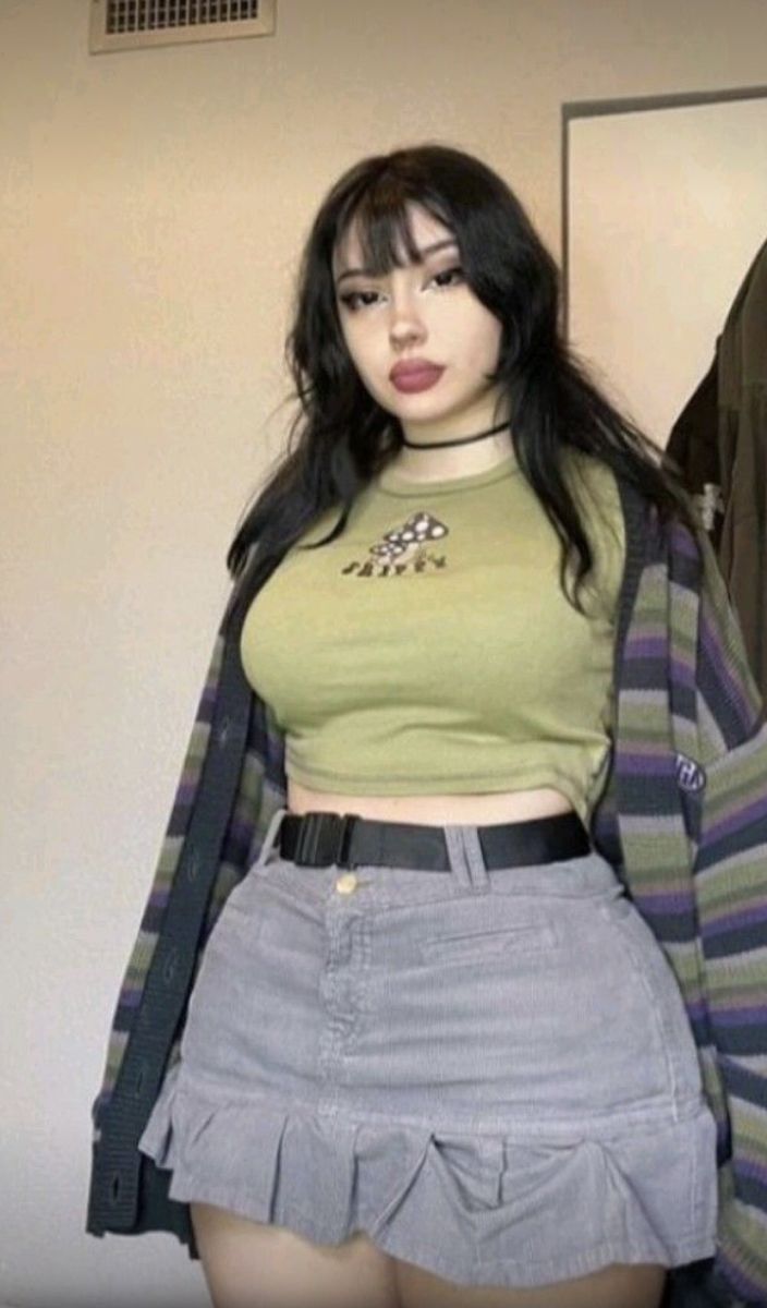 a young woman with long black hair and bangs is posing in a studio wearing a casual outfit.  she has a green crop top with a mushroom embroidered on the front, a gray skirt, and a black cardigan with stripes. she is looking at the camera.