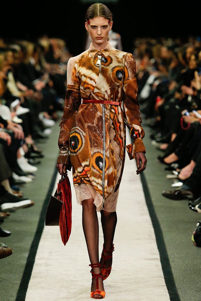 a female fashion model walks the runway in a longsleeved dress with a butterfly print, black tights, and orange heels. she is carrying a red clutch and has her hair pulled back. the runway is black and green, and there are people sitting in the audience in the background.