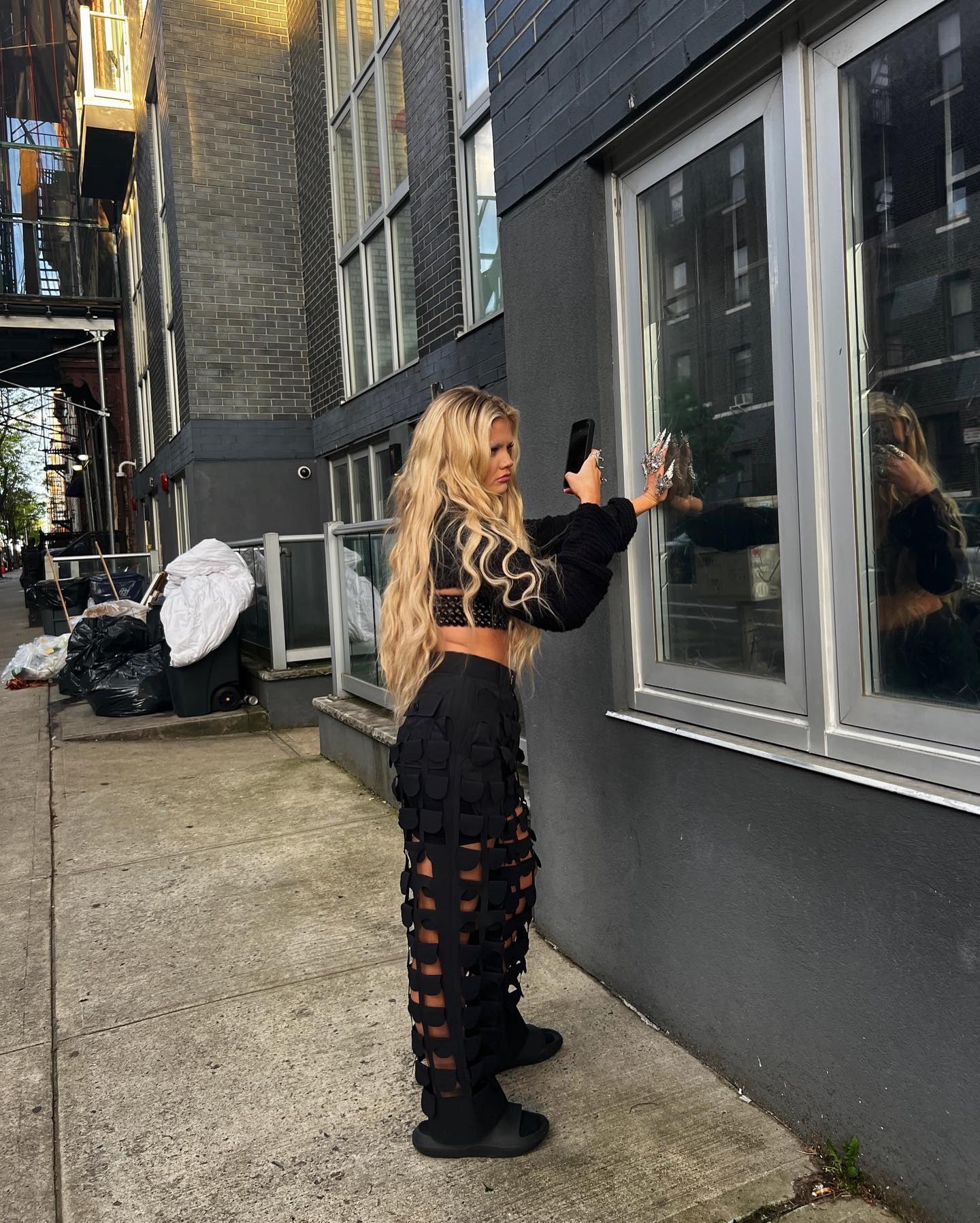 a woman with long blonde hair is taking a selfie in an urban environment. she is standing on a sidewalk next to a building with windows, wearing a black crop top and uniquely styled black trousers. the street scene includes garbage bags nearby. the image captures a casual fashion moment in an urban setting.
