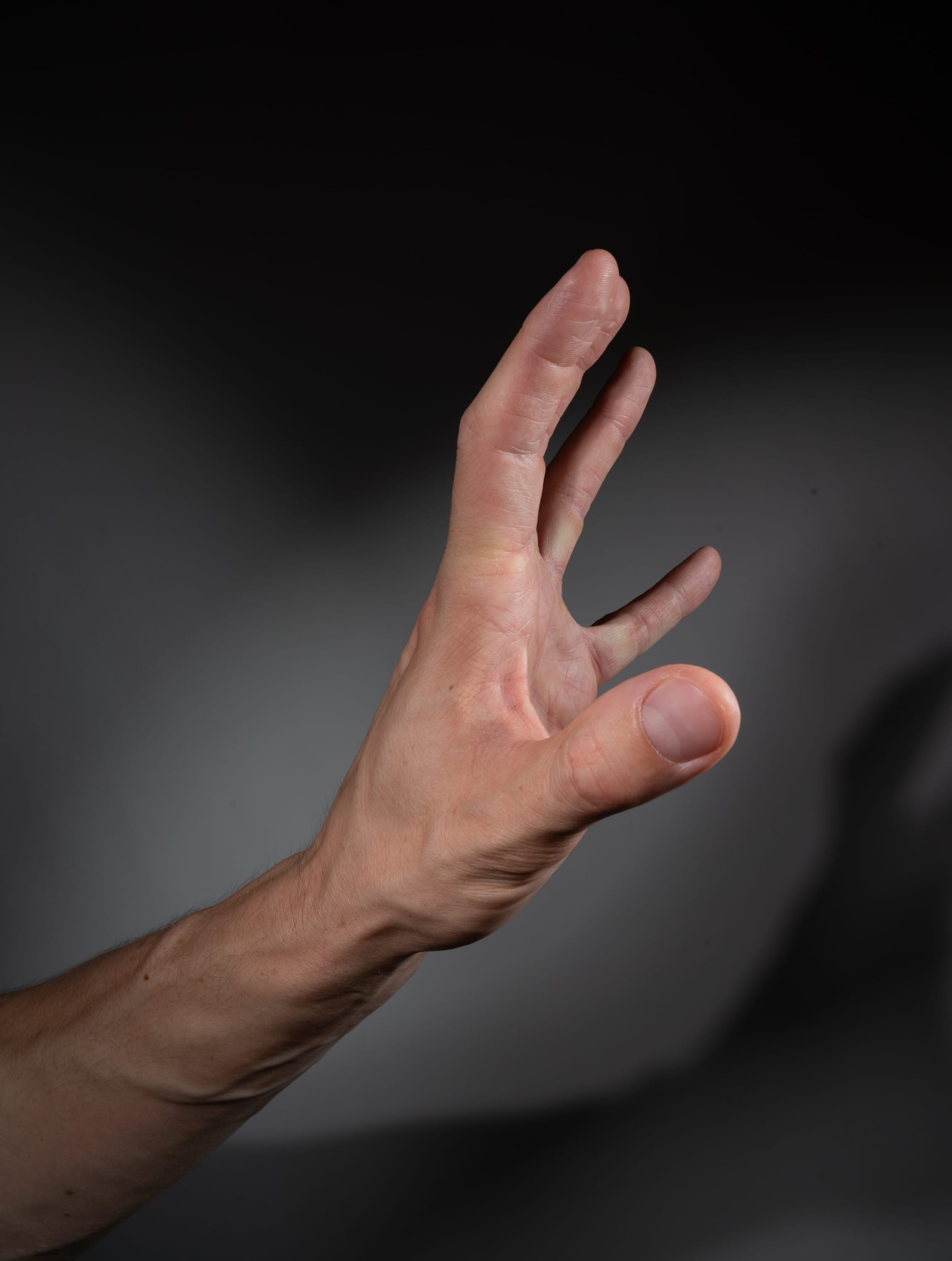 the image shows a man's left hand and forearm reaching upwards against a gray background. the hand is open, with the fingers slightly spread. the lighting is artificial, creating a shadow of the hand on the background. the perspective is a side view.