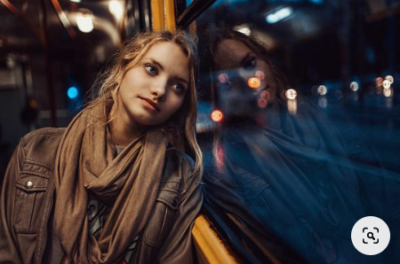 a young woman with blonde hair is sitting on a bus at night, staring pensively out the window. her reflection is visible in the glass, overlaid with the blurry lights of the city passing by. the scene is lit by the dim interior lights of the bus. she is wearing a brown jacket and scarf, giving the image a warm, muted color palette.