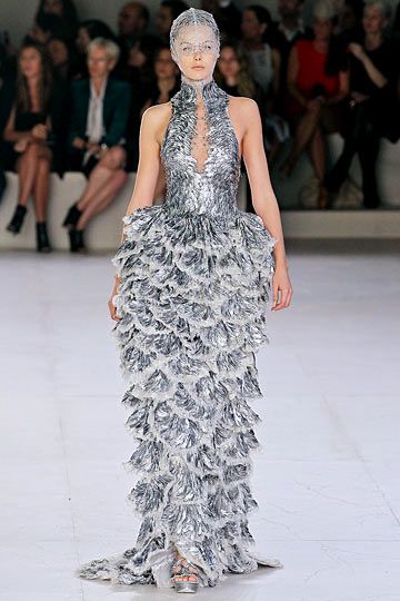 a fashion model walks the runway in a long silver dress. the dress is made up of layers of sequins and feathers, giving it a very textured appearance. the model is wearing a white veil and her hair is styled in a sleek updo. she is posing for the cameras with her hands on her hips. the background is a white runway with a blurred audience. the overall look is glamorous and sophisticated.