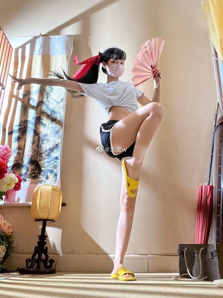 an asian woman wearing a face mask, tshirt, and shorts is standing indoors while posing on one leg. she is holding a pink fan in her right hand and has her left arm extended to the side. she is wearing yellow slides on her feet. the background includes antique furniture and decorations.