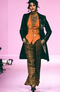 a woman wearing a black leather coat, plaid pants, and an orange vest walks the runway during a fashion show.  the woman is posing for the camera and has a confident expression on her face.  the photo is shot from a slightly low angle, highlighting the model's outfit and the runway setting. the woman's footwear isn't visible in the image.