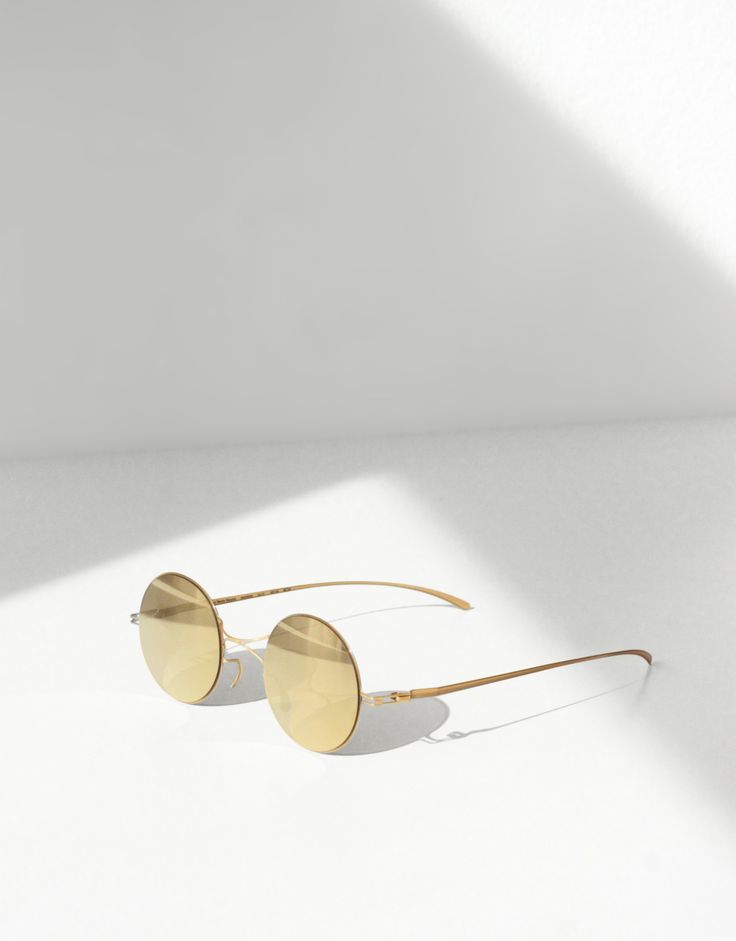a pair of gold, round sunglasses are laying on a white surface. the sunglasses have reflective lenses and a minimalist metal frame. a shadow is cast on the surface behind the sunglasses. the background is a plain white.