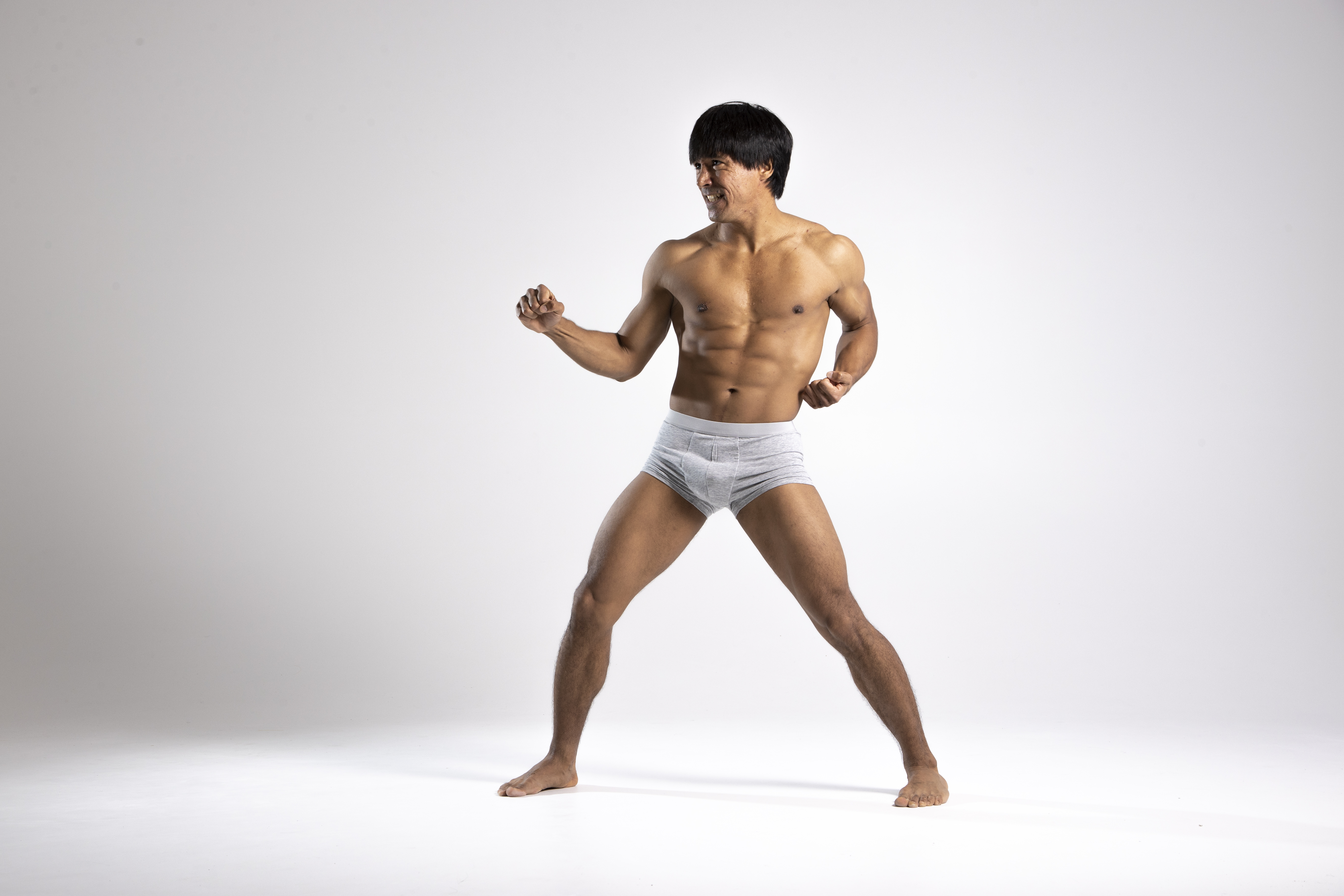 a shirtless man in gray underwear stands on a white background with his legs spread wide and his fists clenched. he has dark hair and appears to be mimicking a martial arts stance, possibly inspired by bruce lee. his toned torso and abs are visible.
