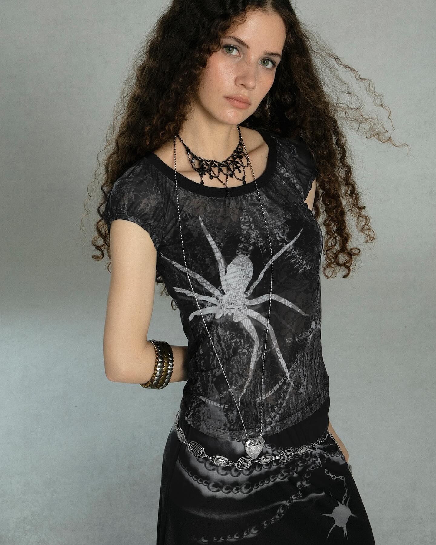 a young woman with long, curly dark hair is posing for a portrait. she is wearing a black and gray sheer top with a spider print, a black choker necklace, several bracelets, and a metal belt with a heartshaped pendant. her skirt is also black and gray with a chain print. she has a serious, slightly intense expression. the background is a plain gray.
