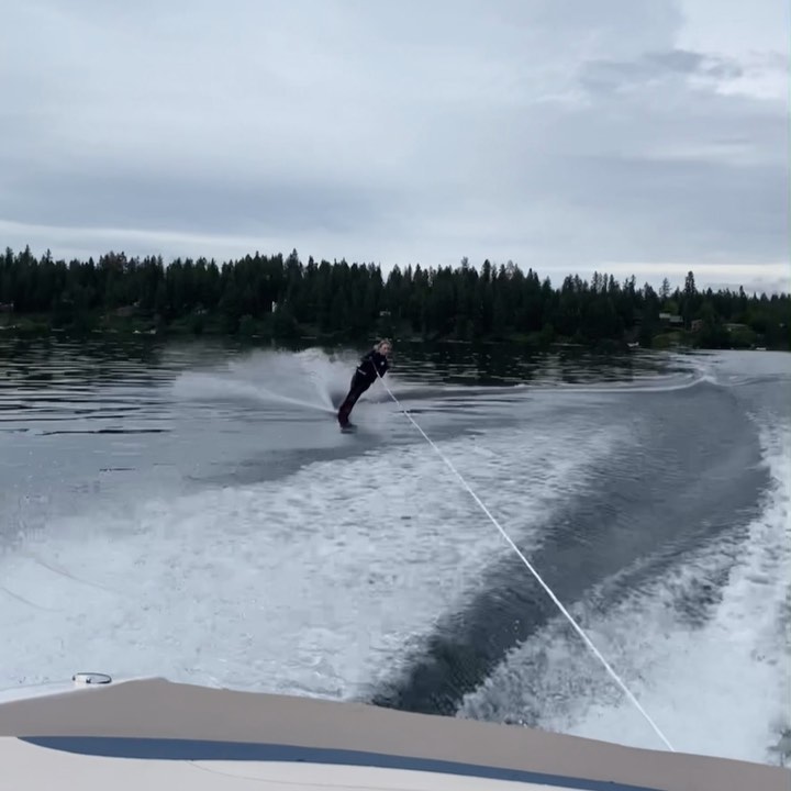 an image shows a person water skiing on a lake. the person is wearing dark clothing and is being towed by a boat, which is partially visible in the foreground. the lake is relatively calm, with some wake behind the skier.  trees line the shoreline in the background, and the sky is mostly cloudy. the overall scene depicts a sunny day with a watersports activity.
