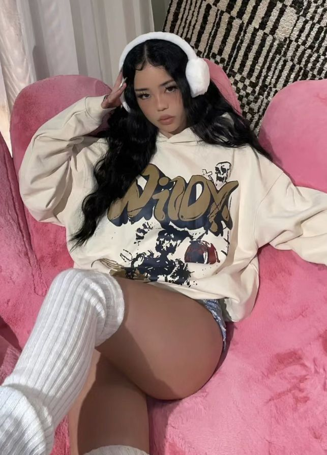 a woman is sitting in a pink chair. she has long, wavy black hair, wearing a light beige hoodie with a graphic design, white fluffy headphones and white kneehigh socks. the background includes patterned fabric behind the chair.