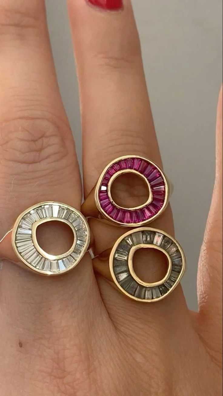 a close up image of a hand wearing three gold rings. the rings are all circular and set with small stones. the ring on the middle finger is set with pink stones, the ring on the index finger is set with clear stones, and the ring on the ring finger is set with dark grey stones. the hand is wearing a clear nail polish.