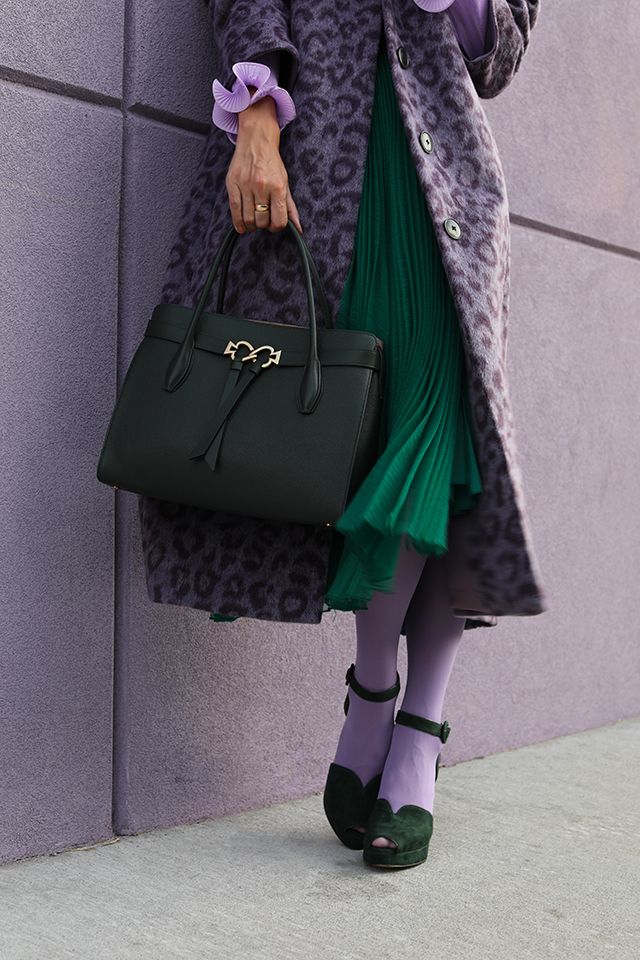 a stylish woman is standing against a purple wall, showcasing a fashionable outfit. she is wearing a purple leopard print coat over a vibrant green pleated skirt, paired with lavender tights and dark green peeptoe shoes. she carries a black kate spade handbag. the overall style is chic and trendy, perfect for fall or winter. the image captures a candid moment of street style fashion.