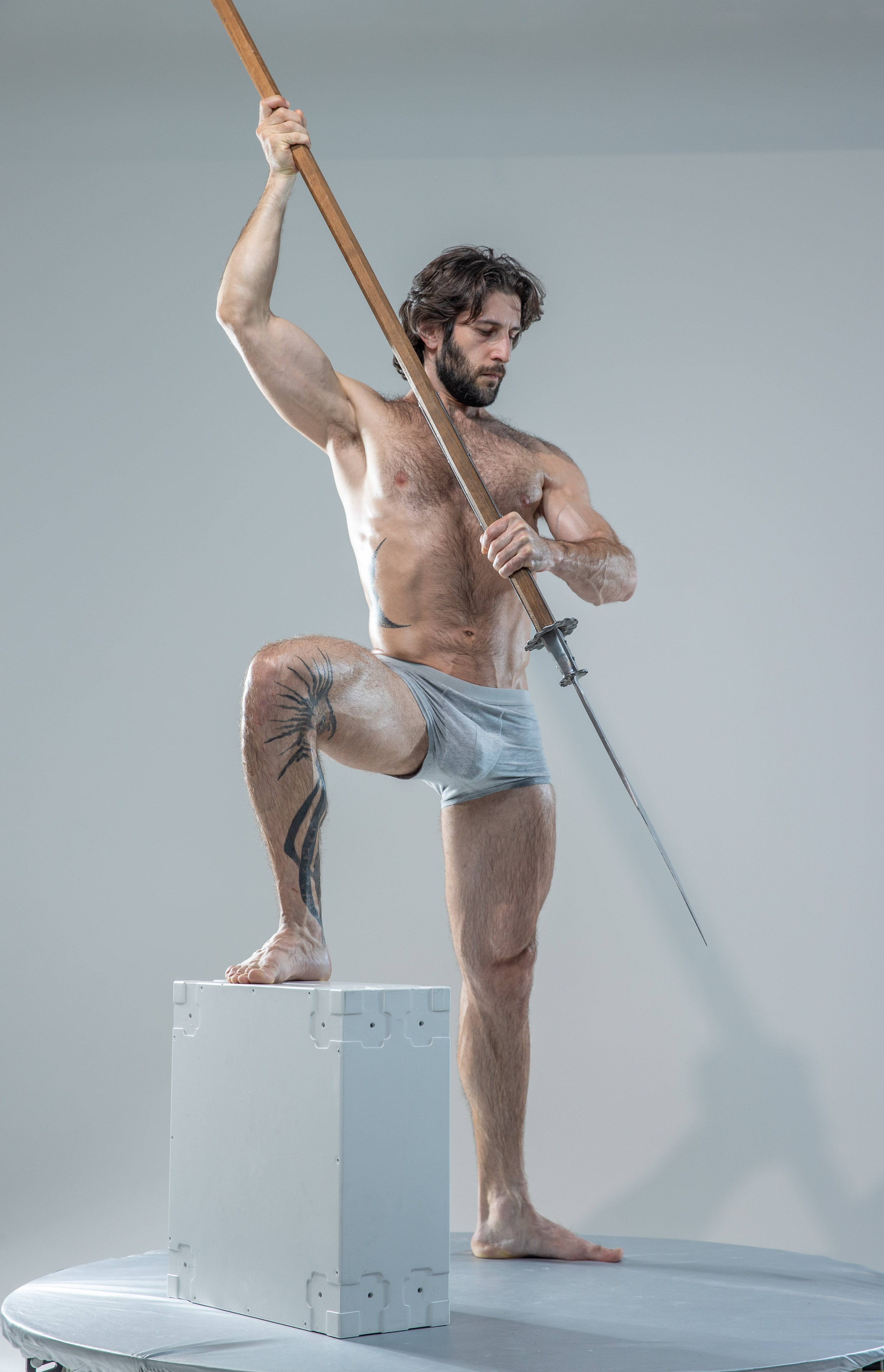 a muscular man with a beard and tattoos is captured in a dynamic pose against a white studio background. he holds a spear aloft, his body tensed with power. he stands on a white platform, his physique illuminated by soft lighting, emphasizing his strength and athleticism. his underwear and the spear provide the only pops of color against the neutral backdrop.