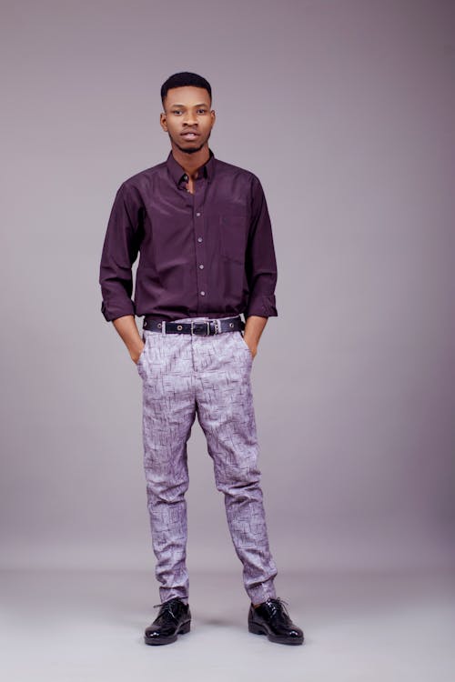 a fullbody studio shot of a young, stylish african american man standing confidently against a grey background. he is wearing a dark purple longsleeve shirt, patterned grey trousers, a black belt, and black dress shoes. his hands are casually in his pockets. the image exudes a blend of formal and casual sophistication, highlighting the model's style and presence.