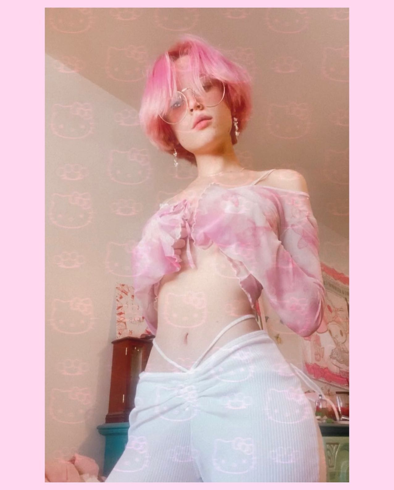 the image features a young woman with short, bright pink hair. she's wearing a pastel pink sheer longsleeved crop top and white pants with a drawstring waist and a subtle hello kitty print. the pants are gathered at the waist. she's also wearing round glasses. the background is a light pink with a repeated hello kitty pattern. the overall aesthetic is very girly and pasteltoned. the photo appears to be taken indoors in a room with some visible furniture in the background.  the woman is posing, looking directly at the camera.