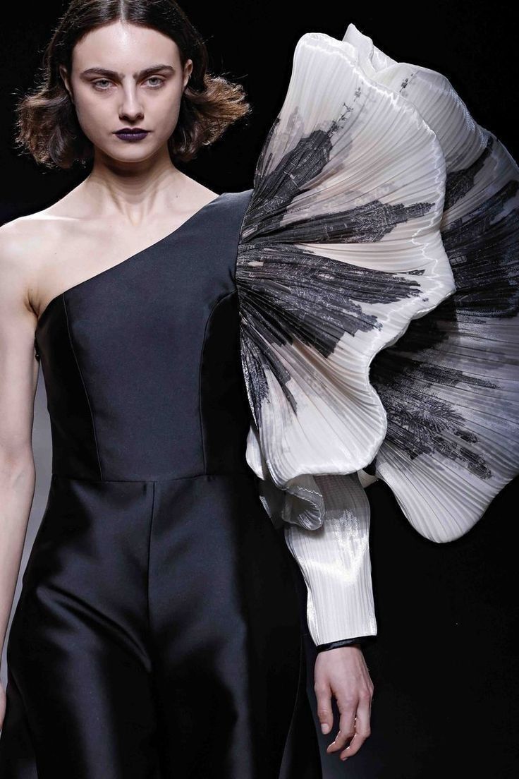 a woman poses on a runway in a striking oneshoulder black dress with a dramatic white sculpted sleeve that resembles a large flower. the sleeve is pleated and features a black and white print, giving the design a futuristic and artistic feel. the model's hair is short and dark, and her makeup is dramatic with black eyeliner and a dark lip.  this is a highfashion ensemble that showcases a bold and innovative approach to design.
