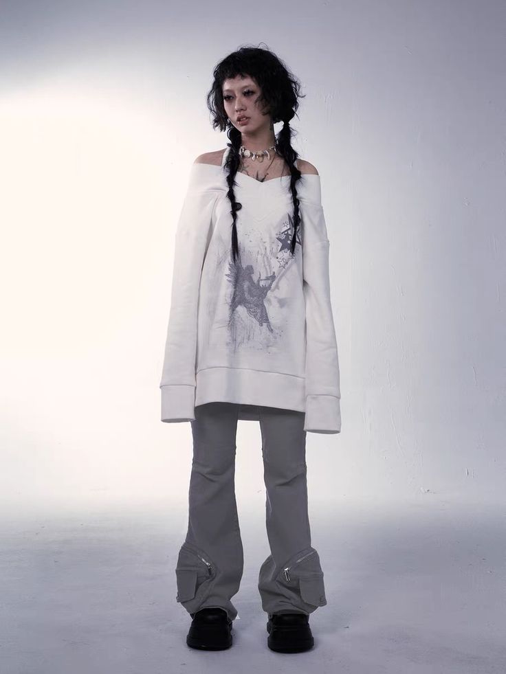 a full shot of a young woman model posing in a studio. she is wearing an offshoulder white sweatshirt with a graphic print, and grey cargo pants. her hair is dark and styled in pigtails. she is wearing a necklace and her expression is neutral. the background is white, and the overall style is edgy and alternative.