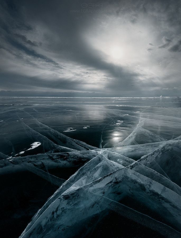 an aerial view of a frozen lake with large cracks and shards of ice in the foreground. the background shows a cloudy sky with the sun shining through. the ice is translucent, and the texture is very prominent.