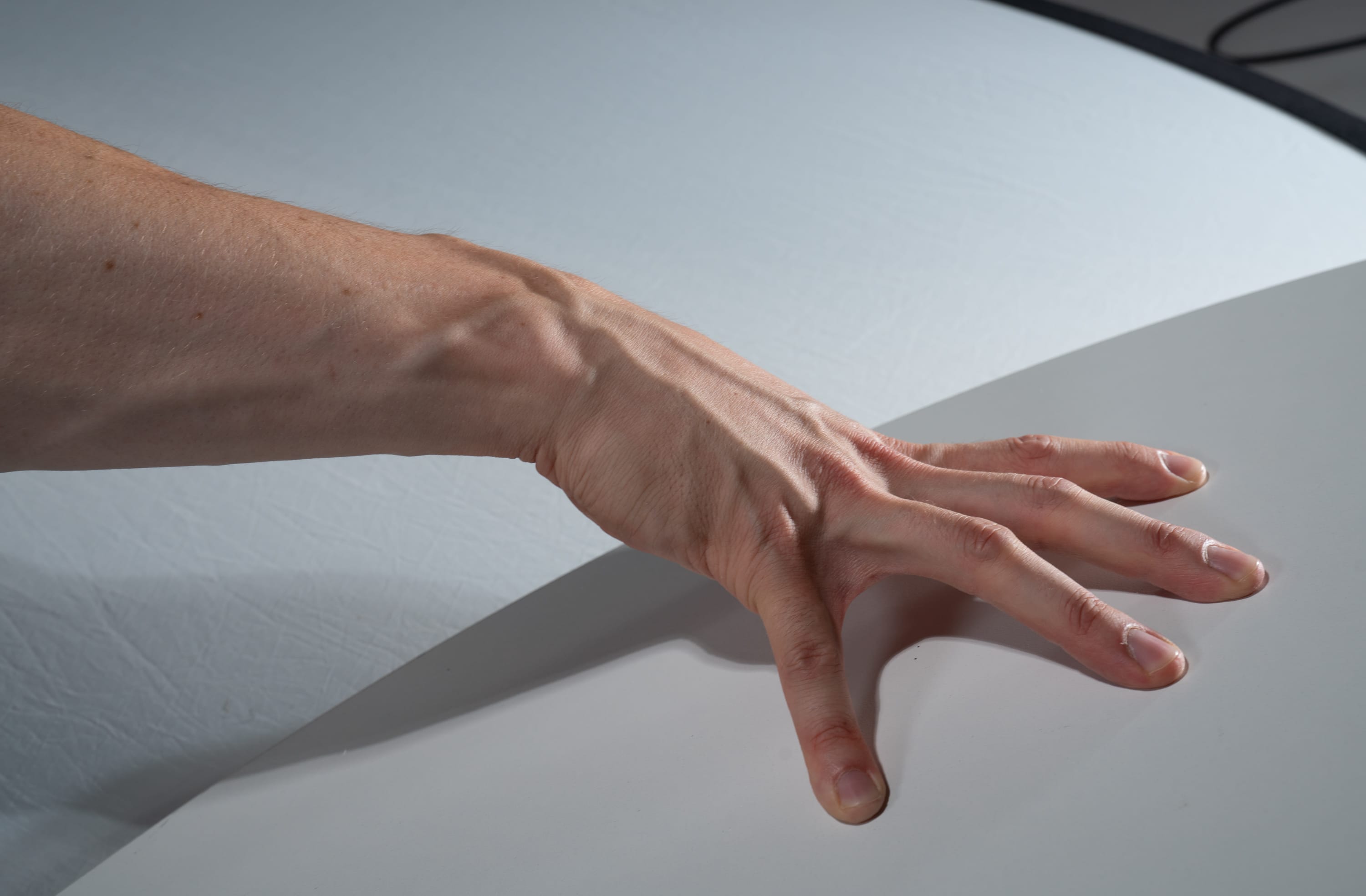 the image shows a closeup view of a person's hand and arm resting on a white surface. the hand is outstretched with the fingers slightly curled. the lighting is soft and diffused, creating a subtle interplay of light and shadow across the skin, highlighting the veins and contours of the hand.