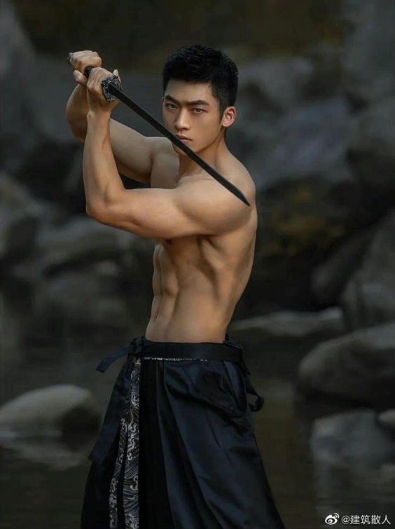 a muscular young asian man with short black hair stands shirtless in a natural environment. he's holding a sword above his head with both hands, seemingly ready to strike. he wears a black skirtlike garment with intricate silver designs. the background features rocks and a hint of water, suggesting a riverbank or similar setting. the lighting is soft and natural, highlighting the man's physique and the texture of the environment.