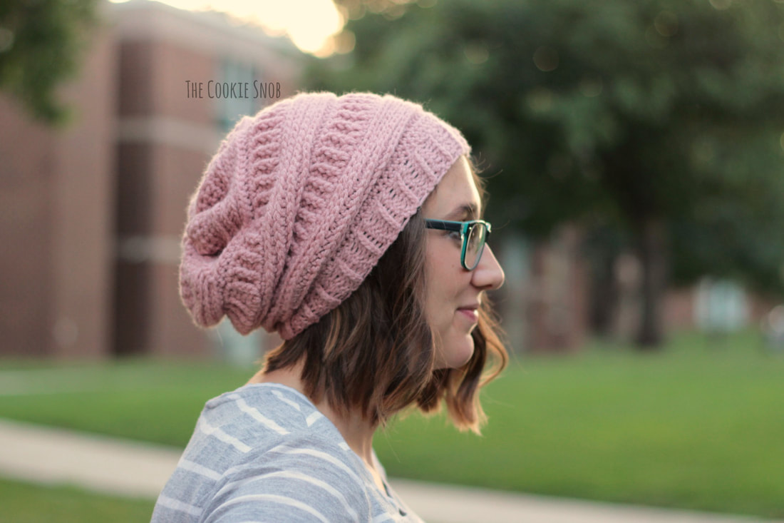 a profile view of a young woman wearing a light pink, slouchy crocheted beanie. the hat is textured with raised cablelike stitches. she's wearing a light gray and white striped shirt. the woman is outdoors, with a blurred background of grass and buildings, suggesting a park or campus setting. the hat design is prominent, indicating a potential advertisement or display of crochet work. the watermark "the cookie snob" is visible in the upper left corner.