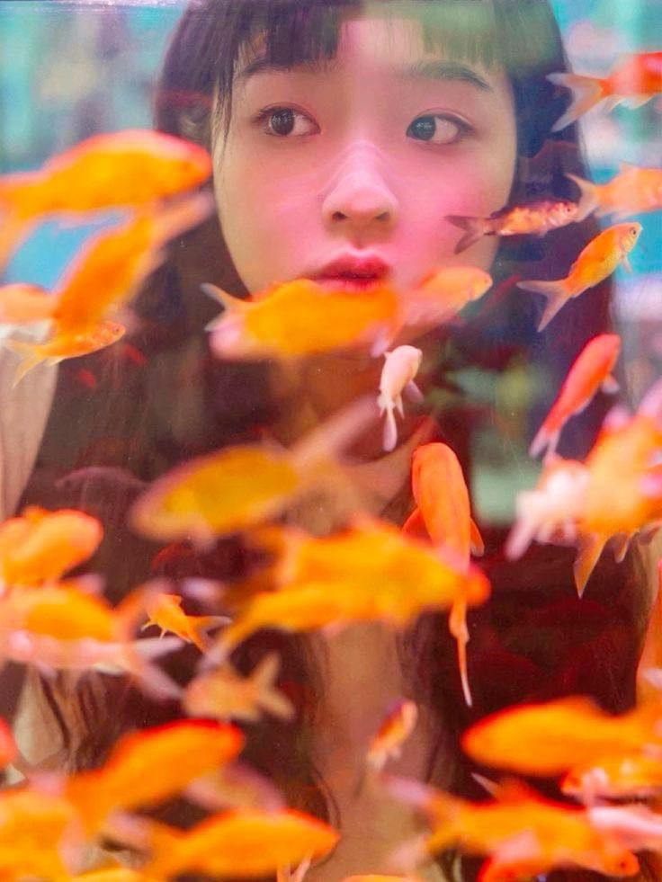 the image is a closeup portrait of a young asian woman with long, dark hair and bangs. she has a calm, almost pensive expression on her face. the woman is positioned in front of an aquarium or fish tank filled with numerous orange and reddish goldfish that are swimming around her. the fish are blurry in places, creating a soft, dreamlike effect. the overall atmosphere is serene and peaceful. the image is artistically styled, with a focus on the contrast between the woman's clear features and the blurred, colorful movement of the fish around her.