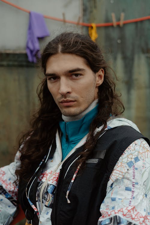 closeup portrait of a young man with long, wavy brown hair. he's wearing a black vest over a white jacket with a maplike print.  the background is a weathered wall with a clothesline and some items hanging on it. the man's expression is serious and intense. the overall style is casual and urban.