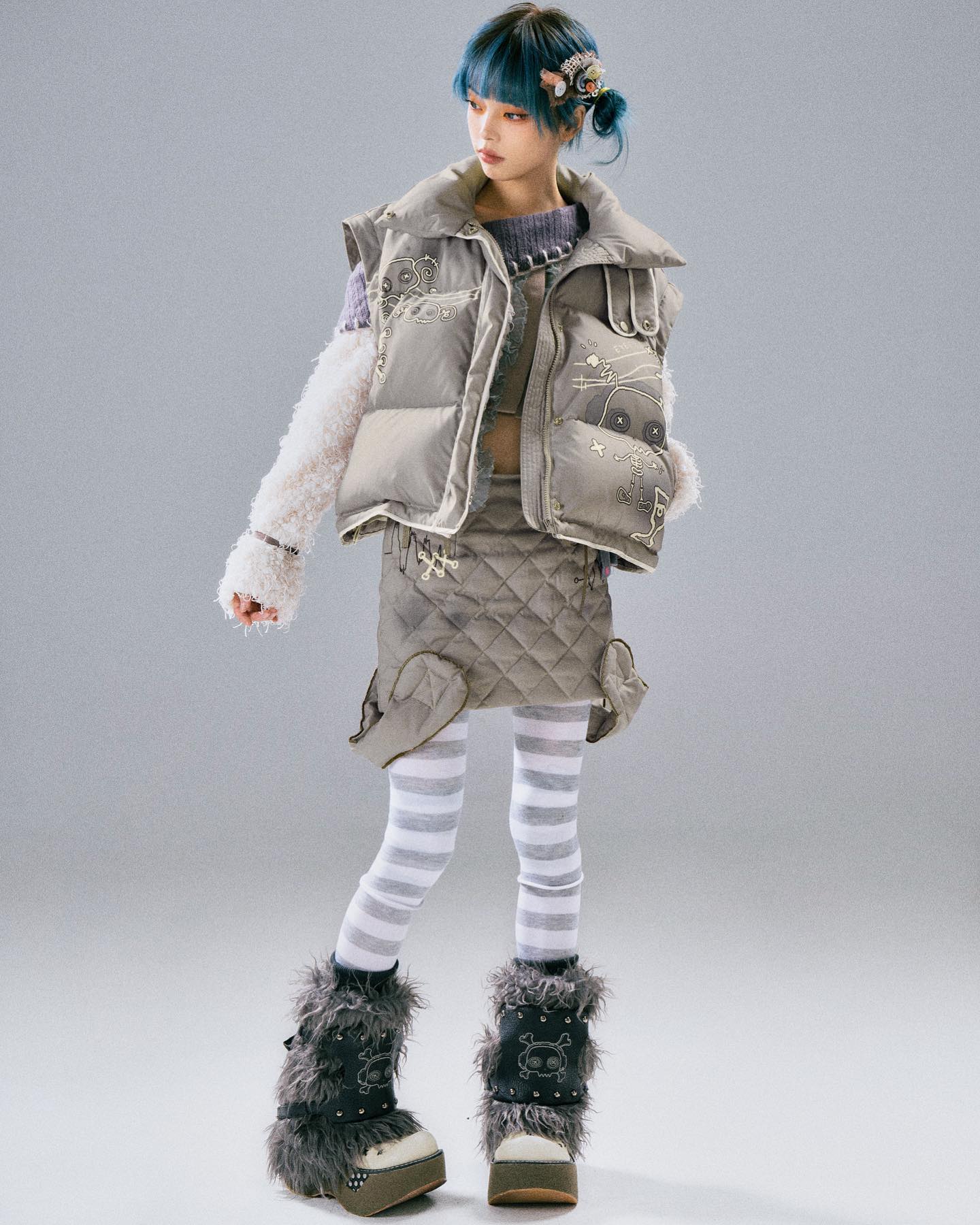 a fullbody studio shot of a young girl model showcasing a stylish winter outfit. she has vibrant blue hair styled in a halfupdo with hair clips. she's wearing a light grey puffer vest with unique embroidery, a matching quilted skirt, white and grey striped leggings, and furry boots with skulllike embellishments. the vest has a relaxed fit and is shown open slightly at the front, revealing a hint of a purple top underneath. the overall aesthetic is a blend of animeinspired fashion and cute, quirky elements. the background is a plain light grey, focusing the viewer's attention on the model and her outfit.