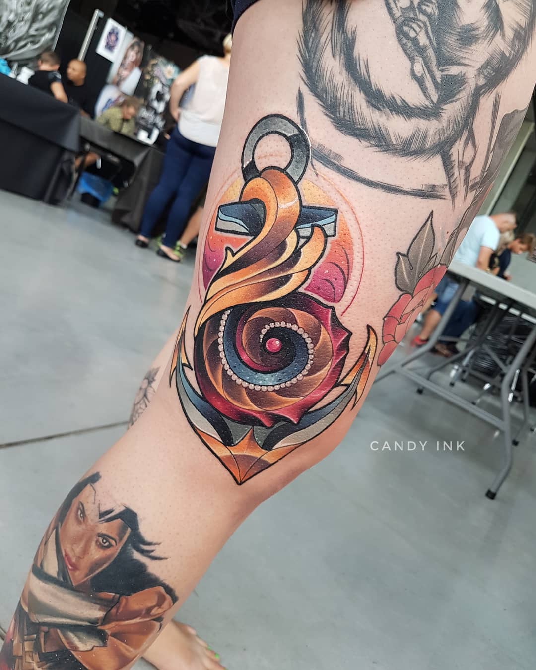 this image showcases a colorful neotraditional anchor tattoo on a woman's upper thigh. the anchor is rendered in a bold, graphic style with a stylized rope design. the color palette is warm and inviting, with shades of orange, red, brown, and yellow dominating the design.  the setting appears to be a tattoo convention, as suggested by the bustling background featuring glimpses of other attendees and booths.