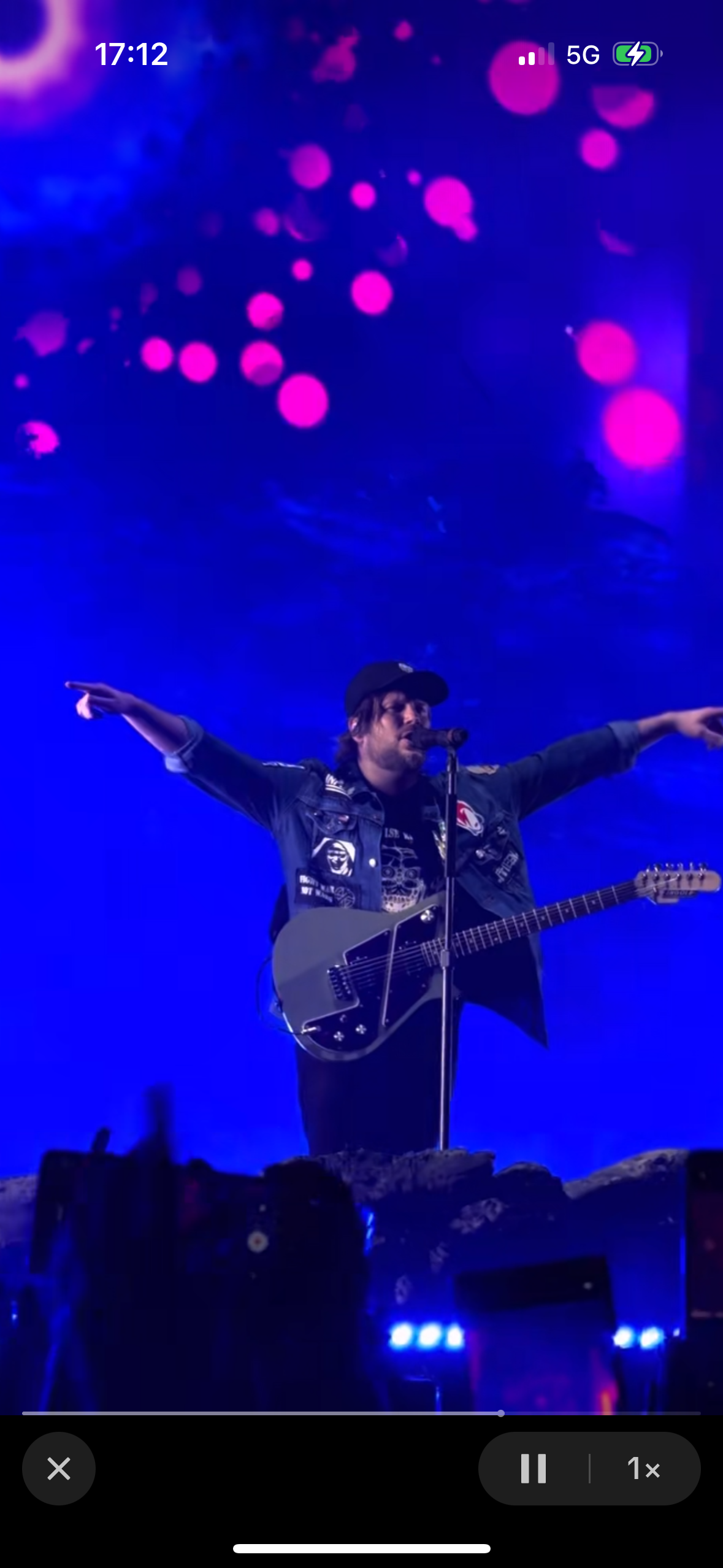 a male musician is performing on stage, playing an electric guitar and singing into a microphone. he is surrounded by bright blue lights and has a crowd of people cheering him on. the background is a blurred image of people and lights. the scene is vibrant and energetic.