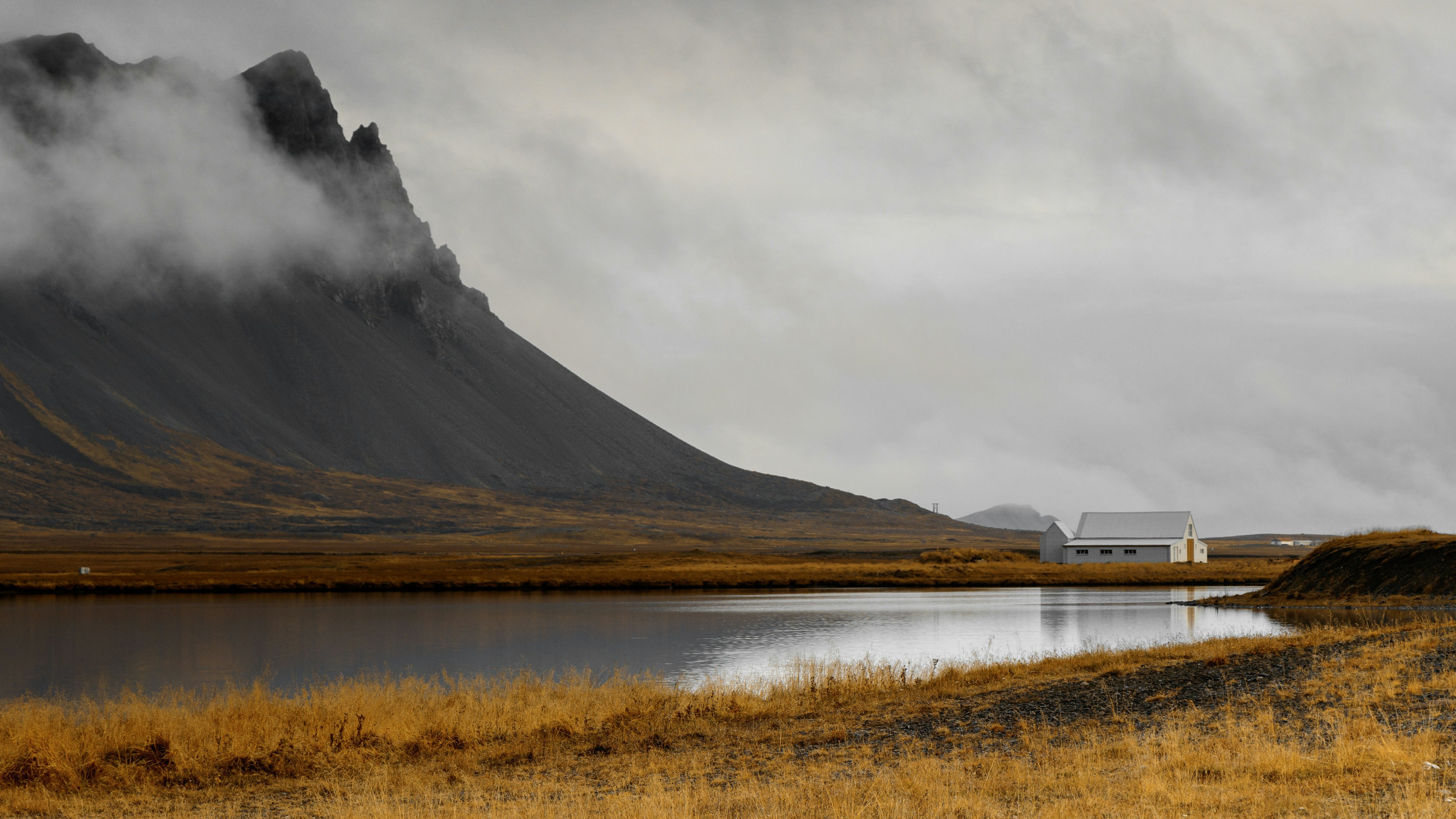 a view of a lake in front of a mountain range, a house is in the distance with mountains and lake reflecting in the water, the sky is cloudy and foggy.  the foreground is a field of dry grass.
