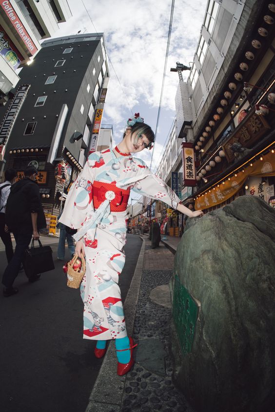 young woman wearing a modern, colorful yukata stands on a city street in japan. she is holding a small wicker basket and leaning on a large green rock. the photo is taken with a wideangle fisheye lens, giving a distorted and dynamic perspective of the urban environment. the sunny day provides natural lighting, illuminating the scene.