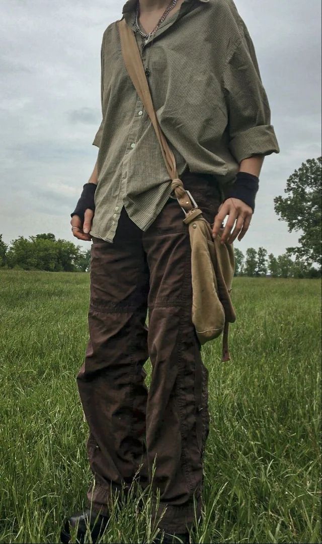 a person stands in a field. the person is wearing baggy brown cargo pants, a light green button up shirt with rolled up sleeves, a crossbody bag, black wrist bands, and a silver chain. the background is a field with tall grass and an overcast sky.